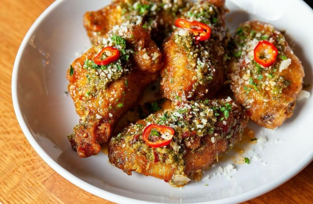 A white plate of crispy wings with herbs and pepper toppings sits on a brown wood table at Station in Pittsburgh.