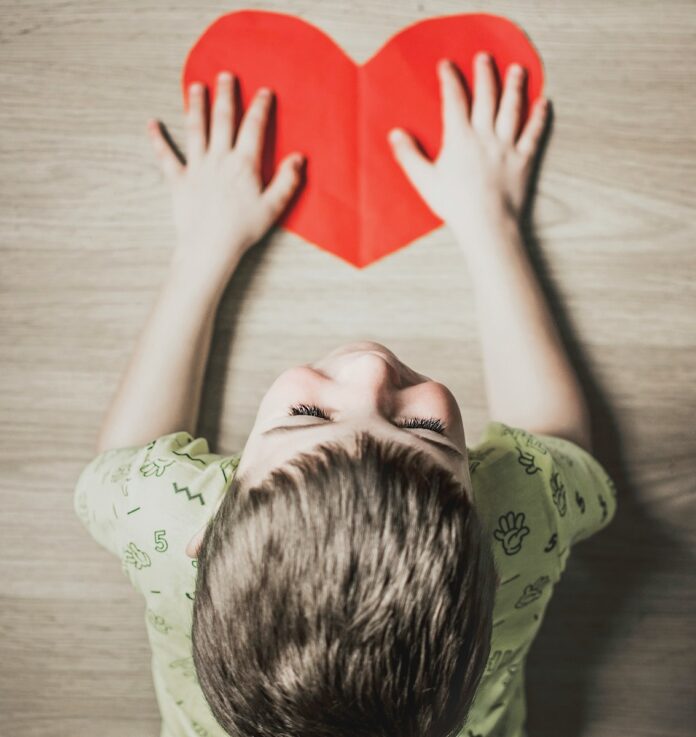 A boy lays on a wood floor with a Valentine's Day red paper heart cut out in front of him.