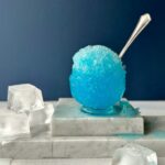A Zamboni, Snow-Cone Cocktail that's a vibrant blue ball of ice in a clear glass cup with a silver spoon sitting on pieces of white marble with large cubes of ice.