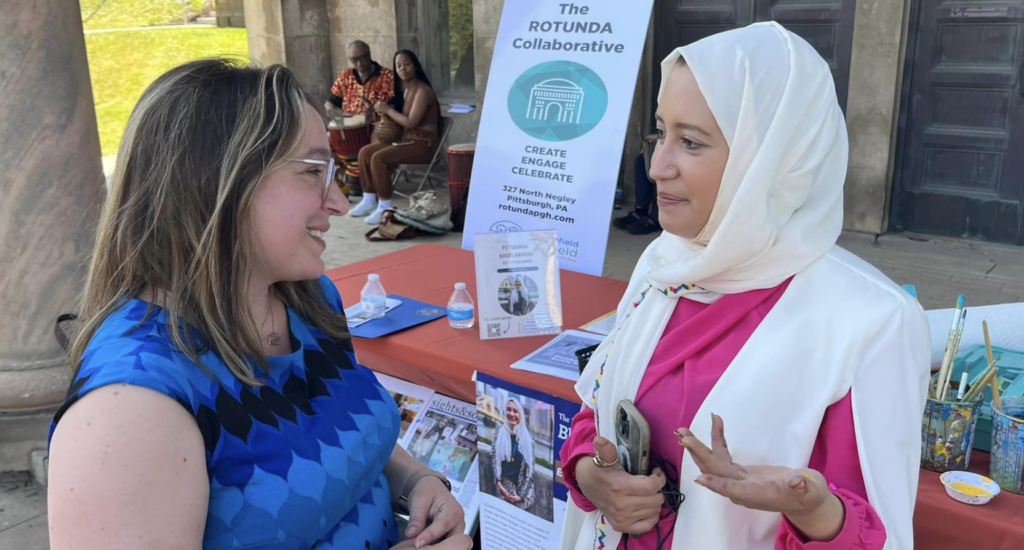 A woman in white a head scarf talks to another woman in blue about the Rotunda Collaborative.