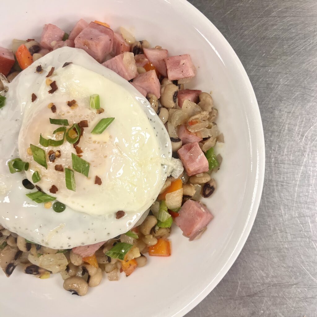 A bowl filled with the Hoppin John; black eyed peas, vegetables, ham, and an egg on top garnished with green herbs.