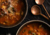 Mark Chutko’s TABLE Favorites Two bowls filled with a dark reddish brown broth with spicy sausage and greens as butter sits to the left and two spoons sit in the top right.