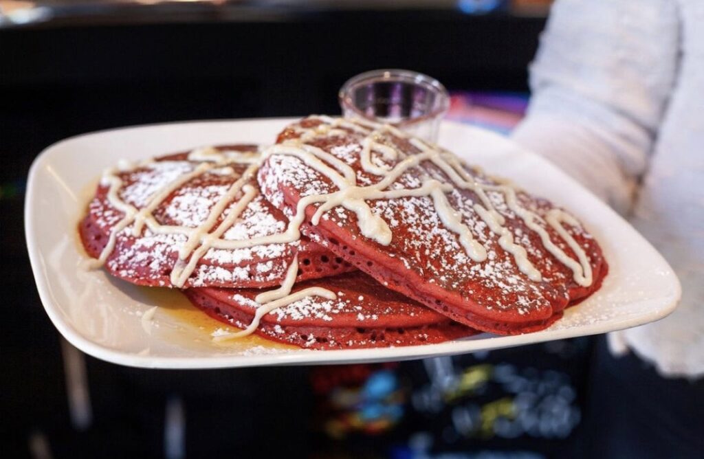 A Valentine's Day special of red velvet pancakes sit on a white plate topped with powdered sugar and cream cheese icing.