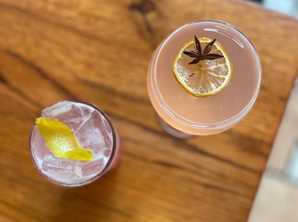 An aerial view of two classes, one filled with a pink lemonade and lemon twist on the right and a martini glass filled with each colored liquid, a lemon, and an anise star on the right. Both drinks sit at a table at DiAnoia's Eatery in Pittsburgh.