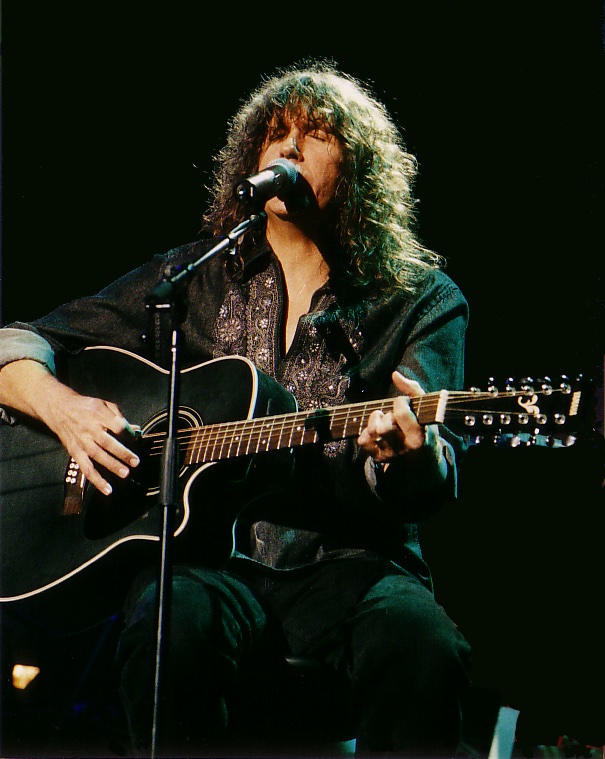 A man with long curly hair plays a black acoustic guitar and sings on stage with the Pittsburgh Symphony Orchestra.