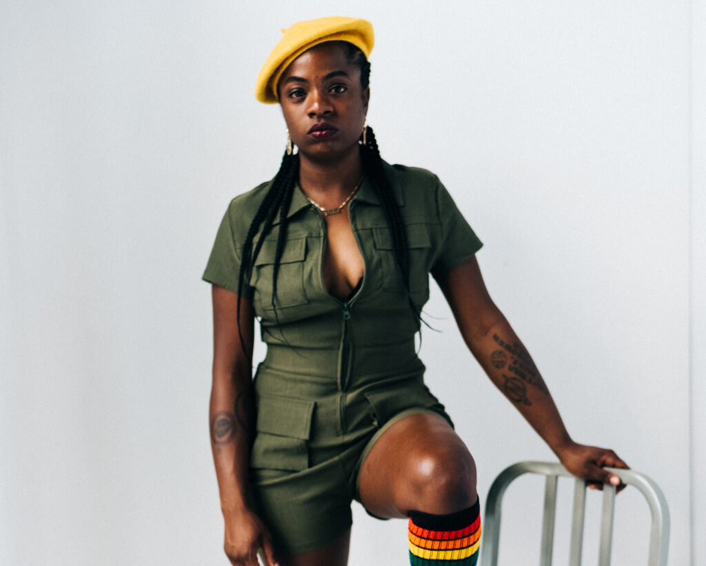 A woman in a green short jumpsuit and yellow beret stands with one food on a white chair in front of a white background.