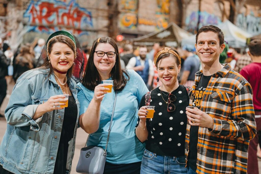 A group of four people stand in a line holding little samples of beer and all smiling at the camera.