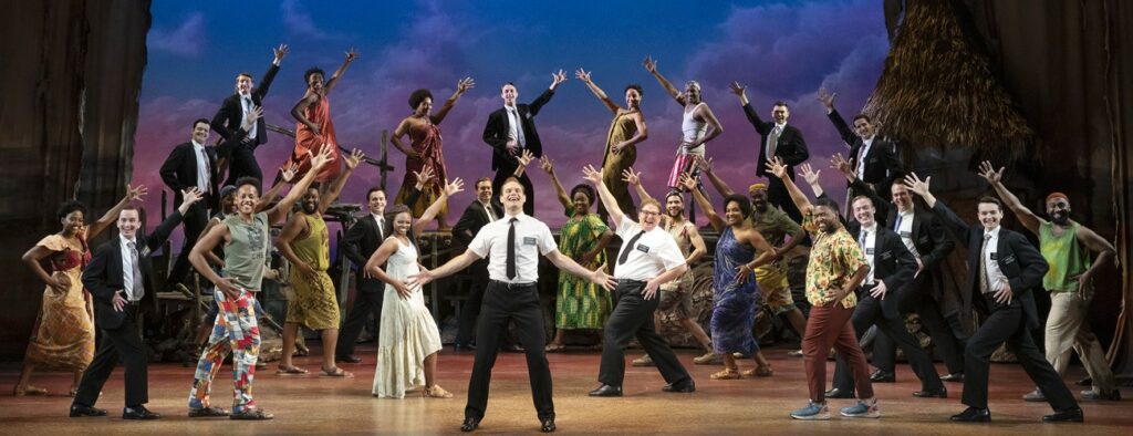 The cast of the book of mormon poses on stage at the end of a big number with Elder Price in a white button up, belting in the middle.