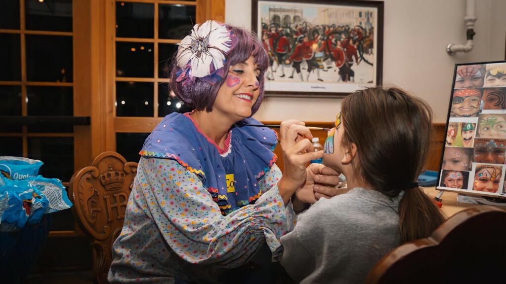 A woman clown with a purple wig paints a blue dot on the face of a little girl with long brown hair.