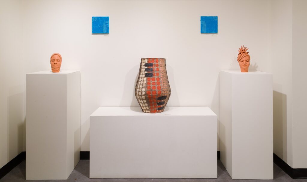 A series of ceramic work including two orange ceramic heads and a ceramic vase on white podiums.