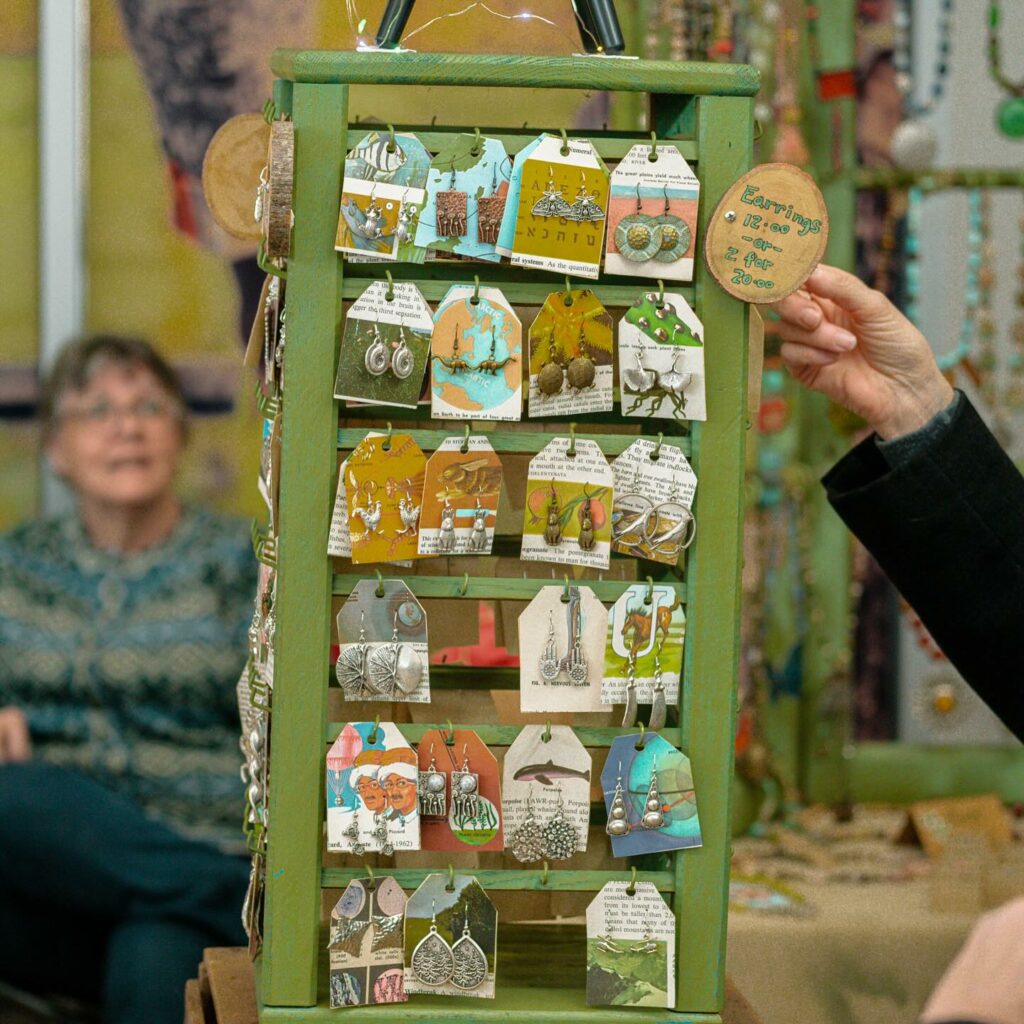 a hand surveys a green, vertical rack of earrings while a woman sits in the background watching at I Made It! Market in Pittsburgh.