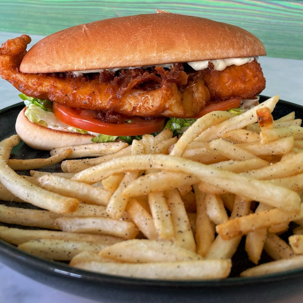 A fish sandwich on a bun with lettuce, tomato, and sauce along with a side of fries sitting in front of it seasoned with pepper.