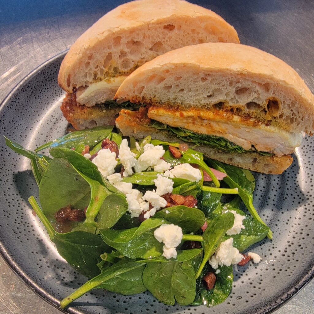 Chicken Romesco sandwich on a ciabatta bun with a side salad featuring cranberries and feta.