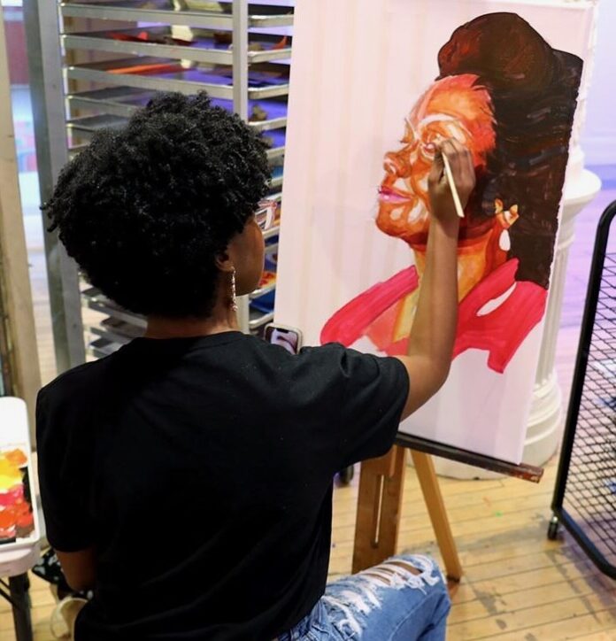 A Black woman sits on a stool painting another Black woman on a canvas as a part of the Children's Museum of Pittsburgh's Black History Month art crawl.