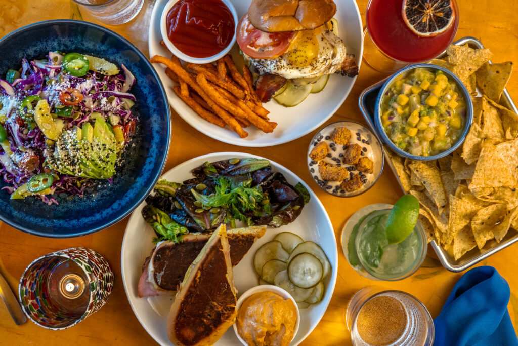 A variety of Cuban inspired dishes such as salads, cuban sandwiches, a burger, and a salsa sit on white and blue plates on a wooden table at Kaya restaurant in Pittsburgh.