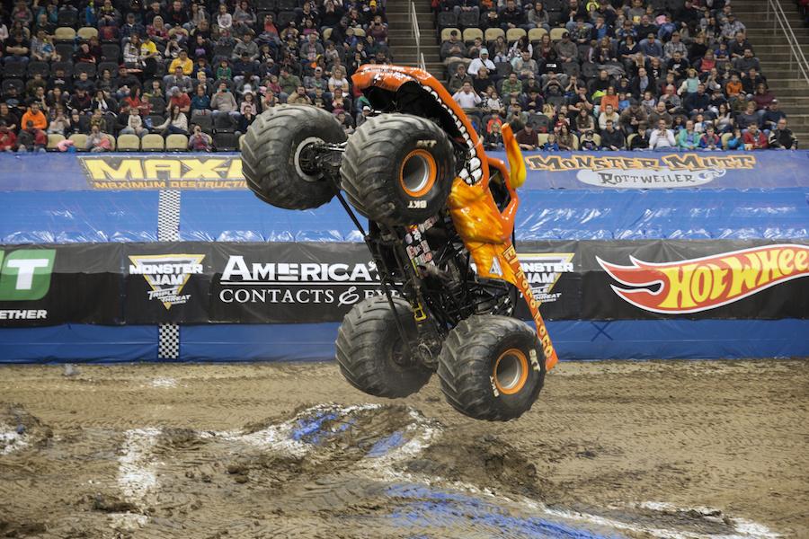 A monster truck in orange colors jumps up vertically over a dirt mound in an arena in Pittsburgh.