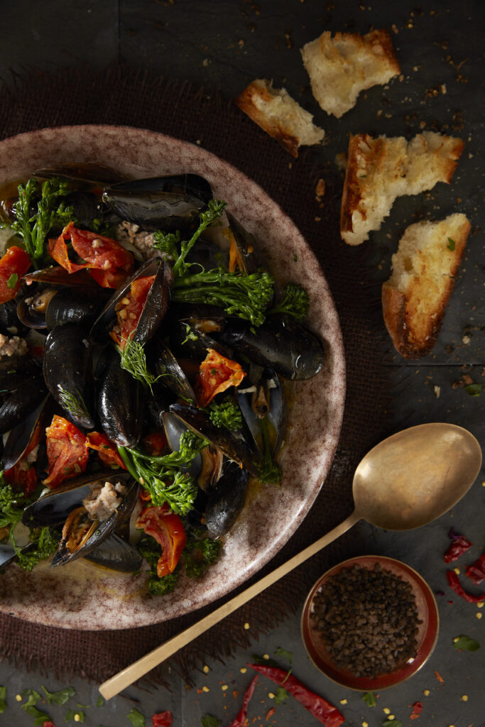 A dish filled with mussels, broccolini, peppers, and more sits on a dark wood table with a crusted baguette and gold spoon to the right.