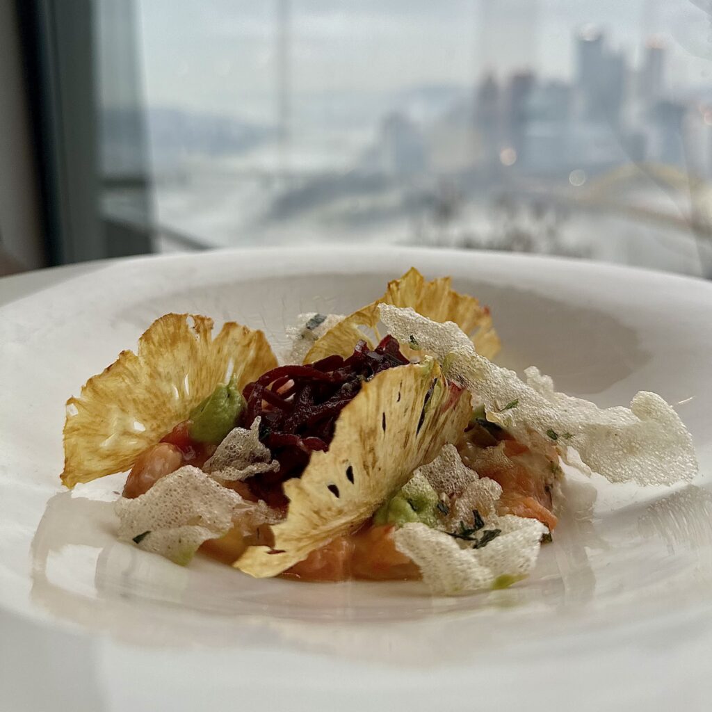 A white plate holds Salmon Ceviche with elegant crisps that look like fans decorating the top all in front of a window showing the Pittsburgh skyline.
