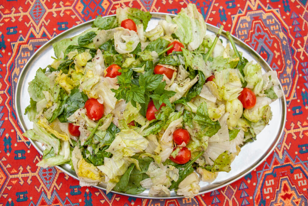 A salad with tomatoes and drenched in dressing sits on a white plate which is on top of a red, Syrian patterned table cloth.