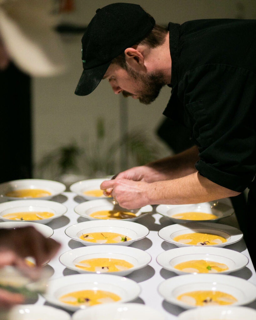 A man in a black hat and shirt adds the final touches to an array of white bowls holding a yellow soup inside.
