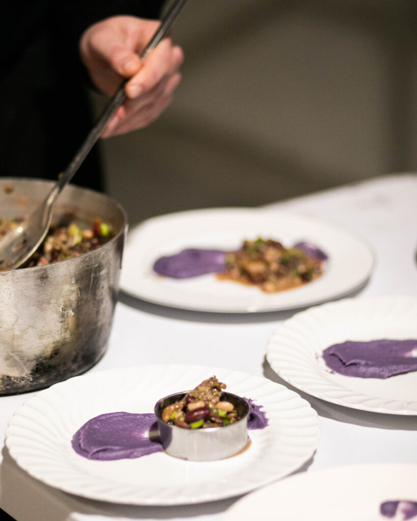A chef plates a purple puree and bundle of stuffing onto three white plates from a silver pot for the famer x baker winter favorites.