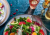 10 Swimsuit Salad Recipes A plate with a salad on it has plenty of kale and citrus fruits like orange slices with drinks in the top right corner of the photo and more plates and forks to the left.