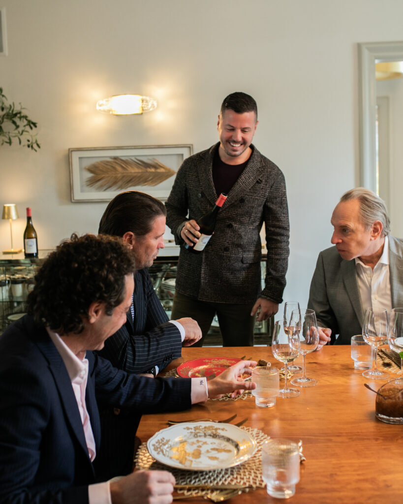 A man in a sweater offers wine to three men sitting at the set table in suits.