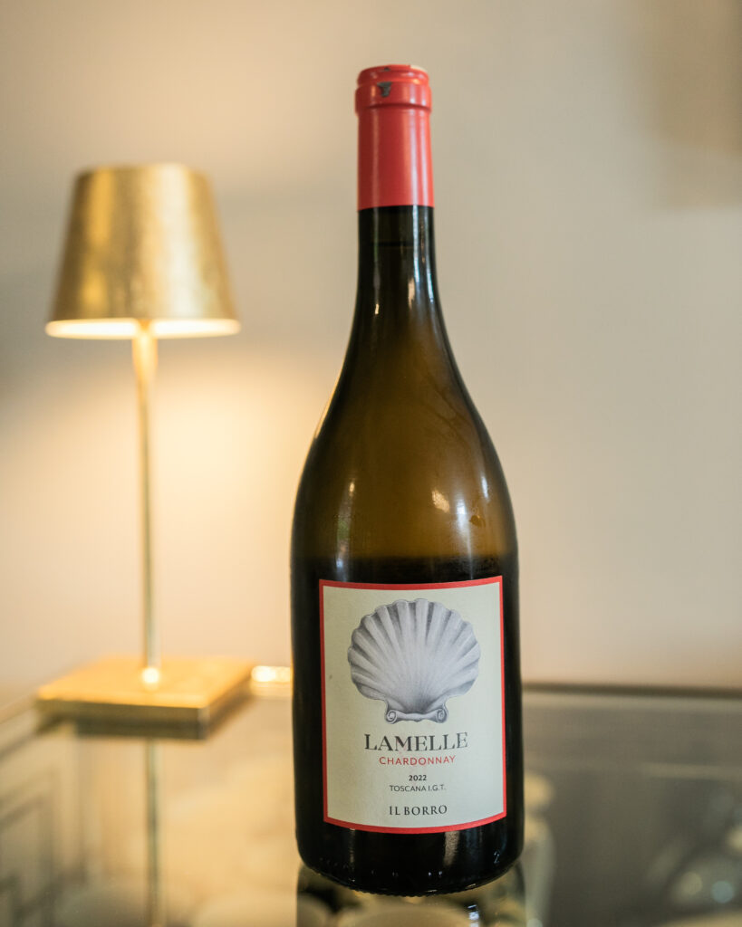 A dark bottle of wine with a blue shell on the label and red cap sits on a dark table with a small golden lamp in the background.