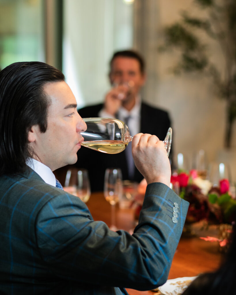 A man sips a light colored wine in a glass while another man, blurry in the background also drinks from a glass of wine.