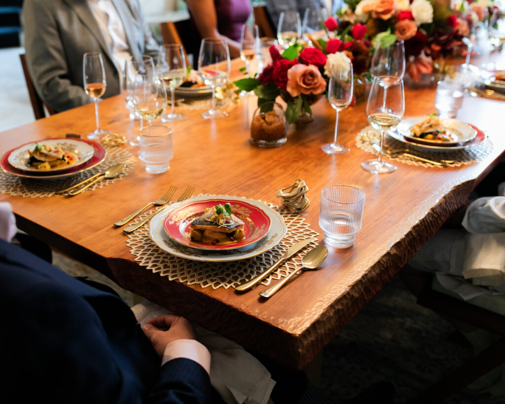 A brown table is set with white and red plates with gold embellishments, topped with food, and plenty of wine glasses and flowers in red and pink around the table at the Il Borro Tuscany Wines tasting.