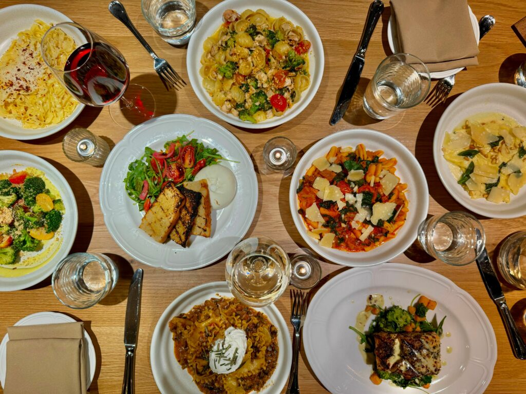 A variety of plates featuring pastas, steak, salads, and other recipes sits on a wooden table with wine glasses dispersed in between the white plates.