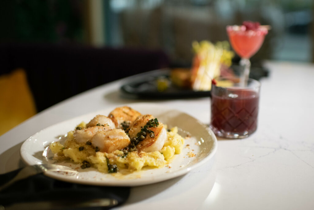 A small white plate holds a shrimp pasta with another darker plate blurred in the background and two red drinks to the right of the dishes.