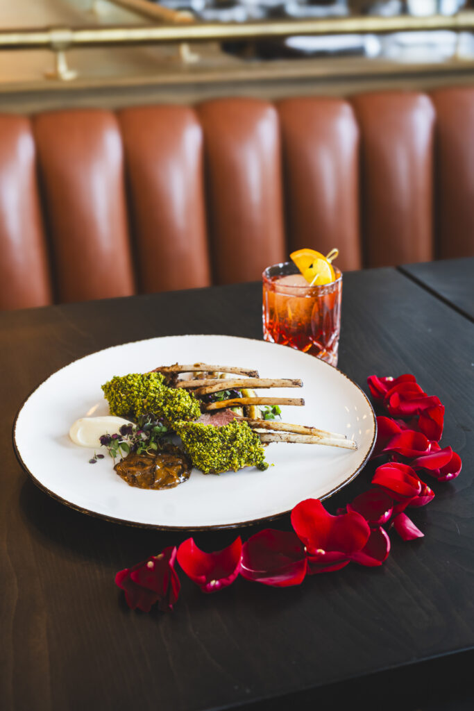 A rack of lamb from Floor 2 sits on a white plate with a sauce beneath the rack and roses surrounding the plate. A cocktail sits behind the plate.