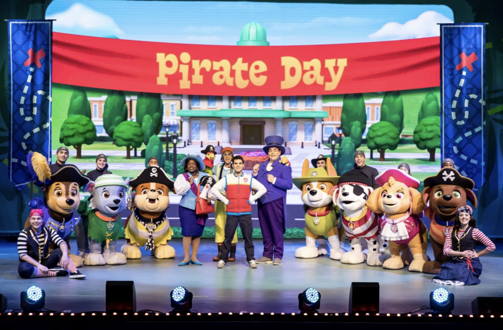 The cast of Paw Patrol Live stands on stage with the mayor and a cartoon looking background banner that says "Pirate Day".