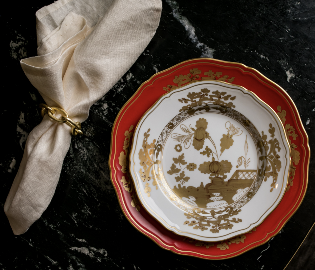 A red plate sits underneath a white plate decorated with gold embellishments, all sitting on a dark marble table with a bundle of silverware wrapped to the left.