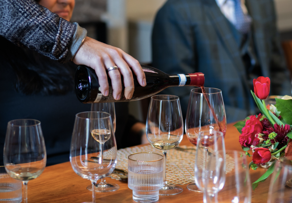 A hand pours a bottle of wine into wine glasses set on a wood table with red roses poking out from the right edge of frame.