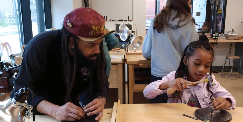 A man in a red baseball cap watches his daughter in a purple shirt hammer a nail into a round piece on a wooden table.