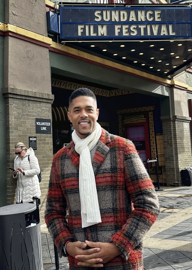 A man dressed in a plaid coat and white scarf stands outside the Sundance Festival sign.
