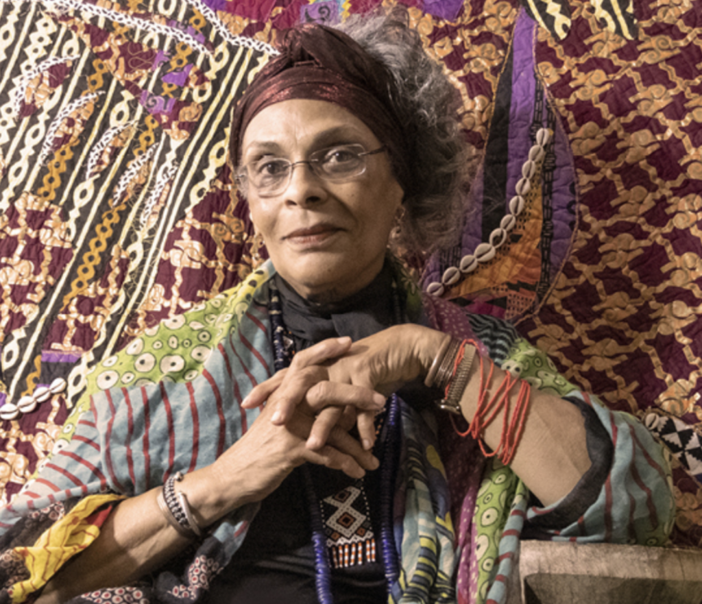 A woman dressed in a headscarf sits against a colorful background with her hands folded in front of her and glasses on her face.