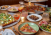 February Menu Updates at Pittsburgh Restaurants A variety of dishes sit on a wooden table holding slices of pizzas, brownies, cannoli's and trays holding whole square and round cut pizzas. Drinking glasses and candles are also dispersed throughout.