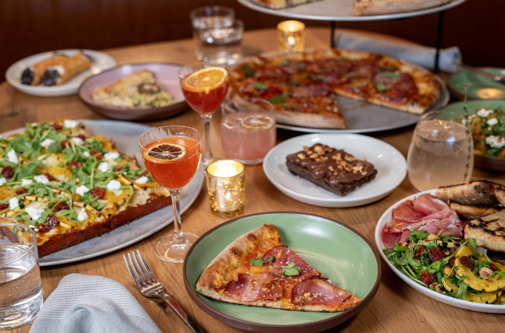 A variety of dishes sit on a wooden table holding slices of pizzas, brownies, cannoli's and trays holding whole square and round cut pizzas. Drinking glasses and candles are also dispersed throughout.