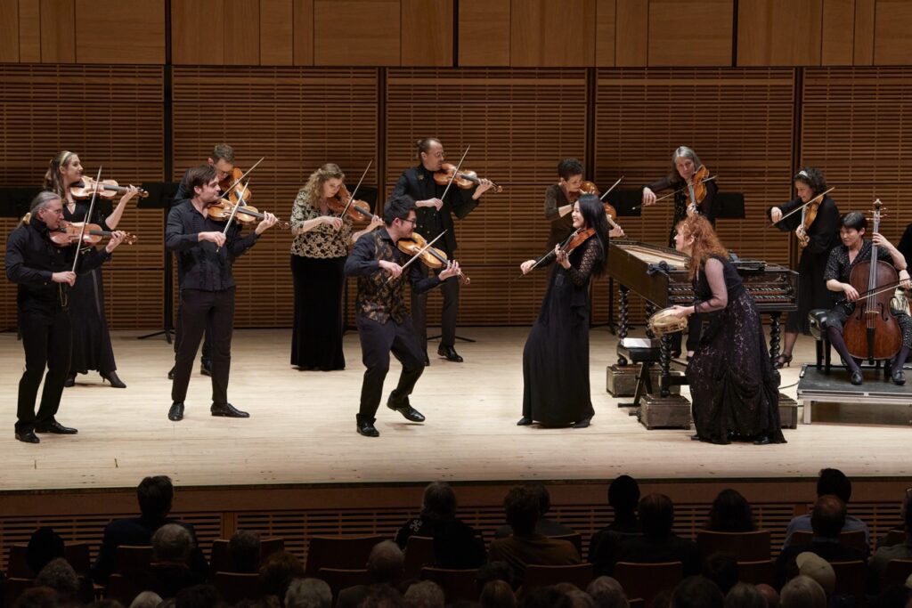 A bunch of violinists in black dress play on a stage with a cello accompanist nearby,