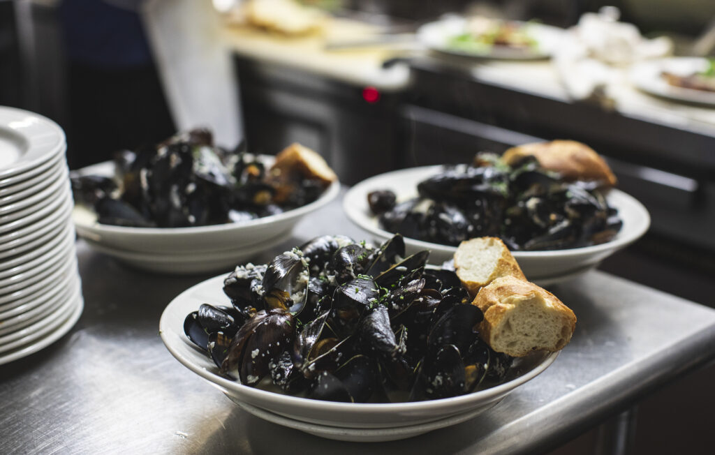 Plates of mussels accompanied by bread sit on a table top in a kitchen waiting to be taken to customers.