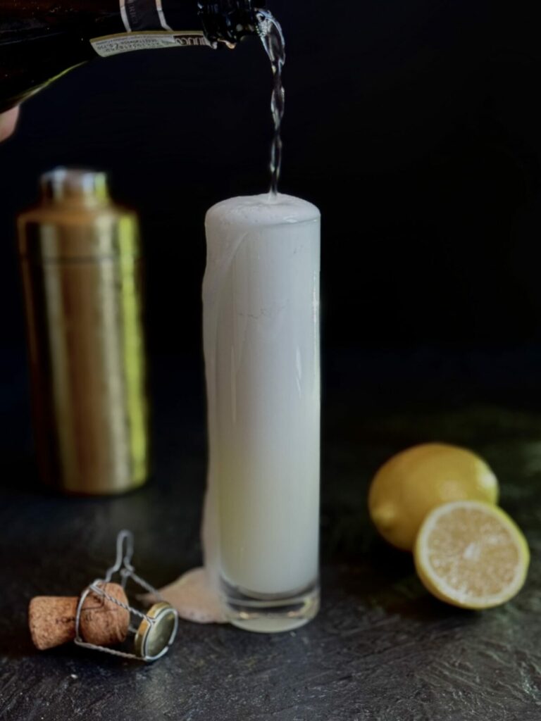 A tall, thin highball glass with a cocktail topped with champagne on a black background with a gold cocktail shaker, lemon, and the bottle cork as styling elements.