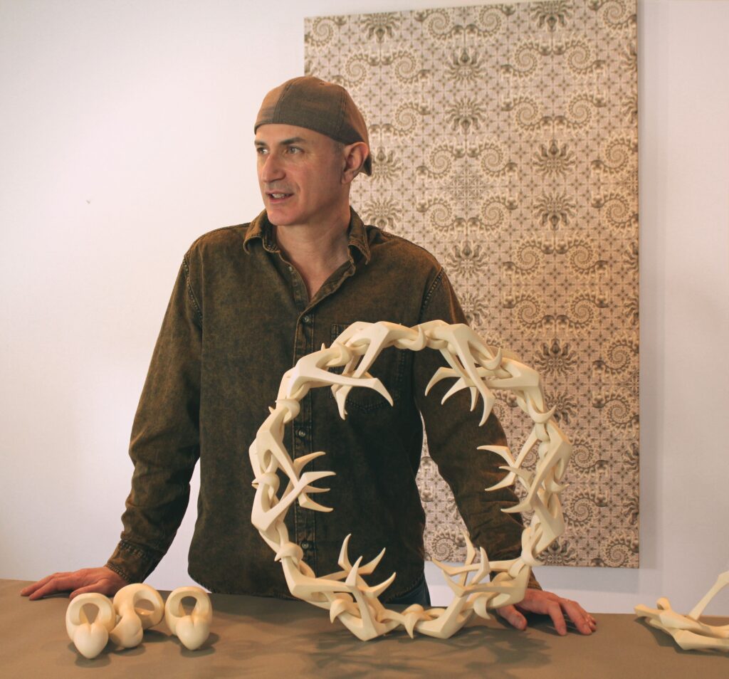 A man in a backwards baseball cap stands behind a table, behind his sculptures.