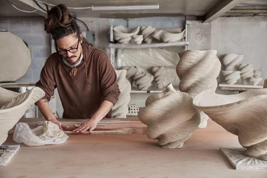 A man works clay at a pottery studio desk surrounded by his creations like swirling vases.