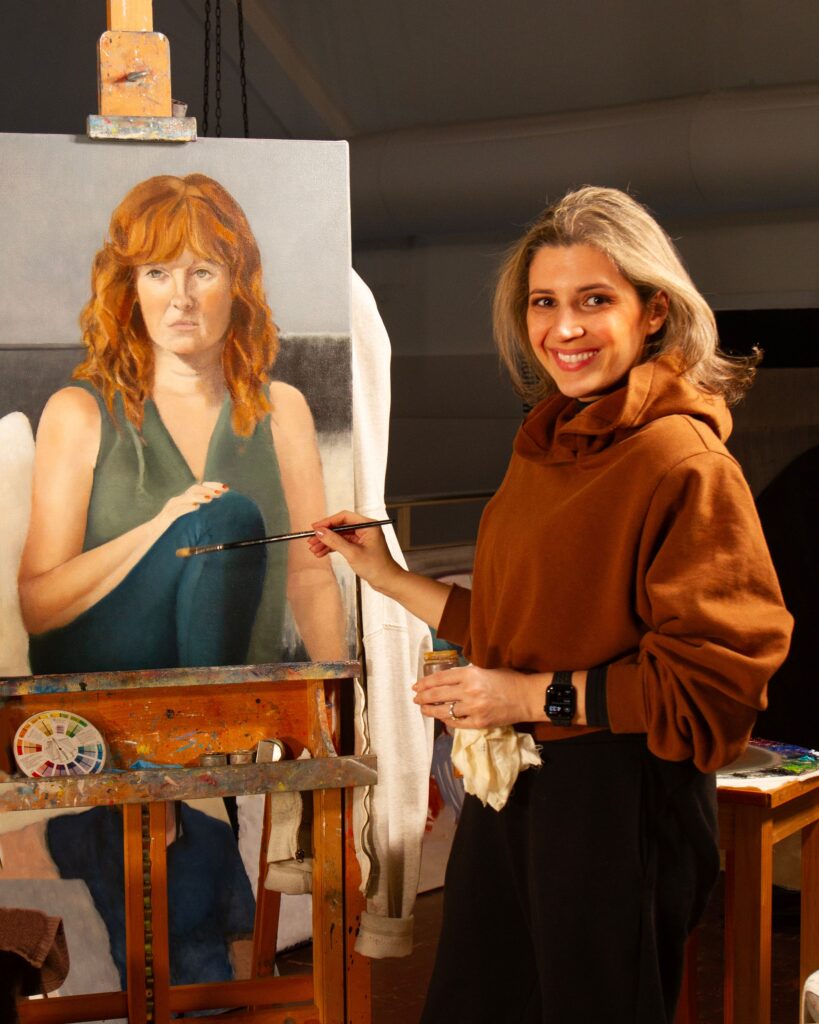 A woman artist in a ochre sweater paints a portrait of a woman at her easel.