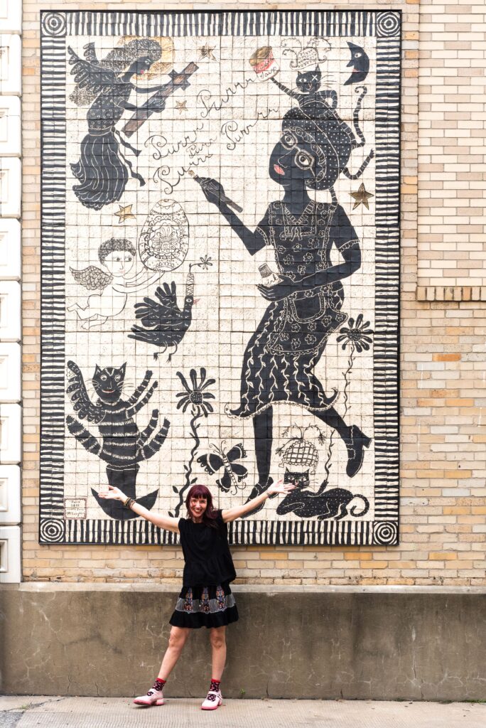 A woman artist stands in front of a tile mural with her hands up in the air presenting the piece.