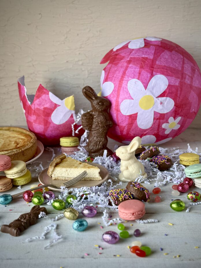 In front of a big, broken pink Easter egg sits various chocolate bunnies, eggs, macarons, ricotta pie, and confetti.