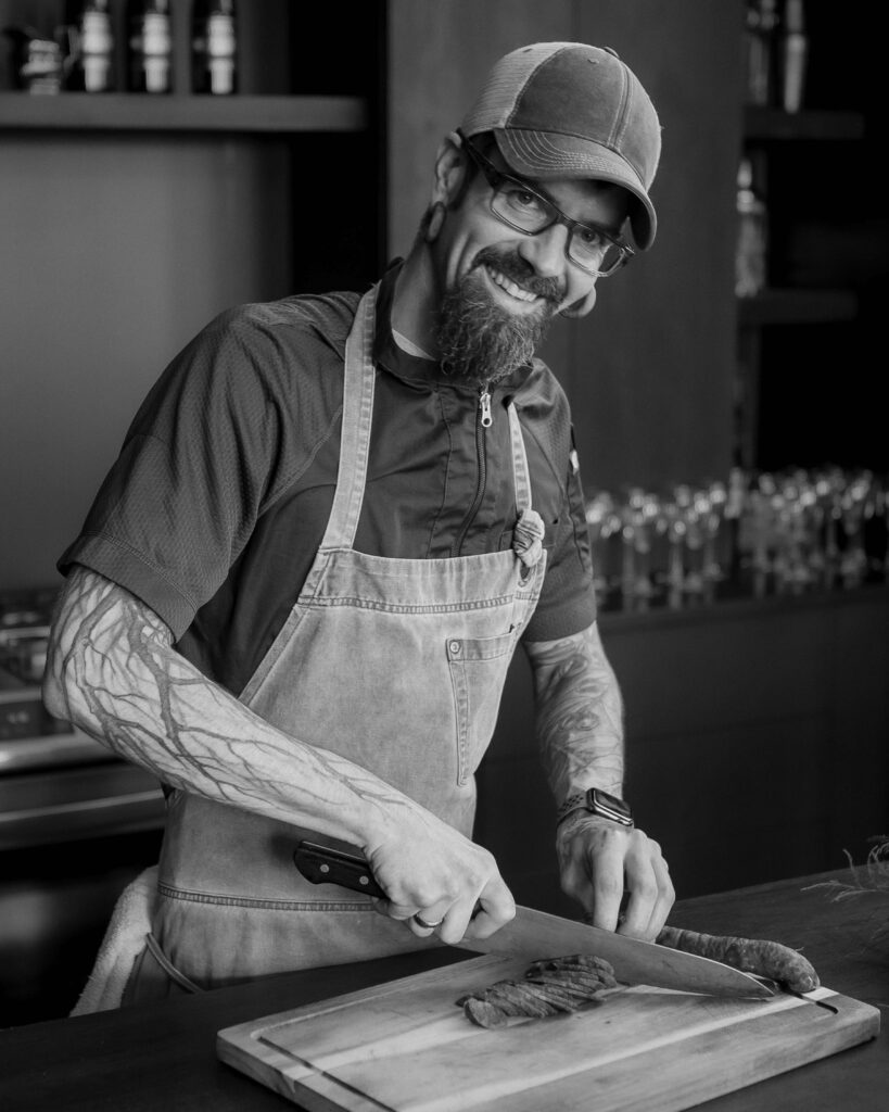 Kevin Hermann cuts a product at a cutting board white smiling at the camera in a baseball hat. The photo is in black and white.